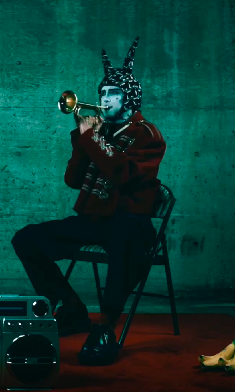 Sir Lucius, seated in a red bandsman's coat and harlequin horned hat, plays a solo trumpet to a boombox with bananas on the floor|320